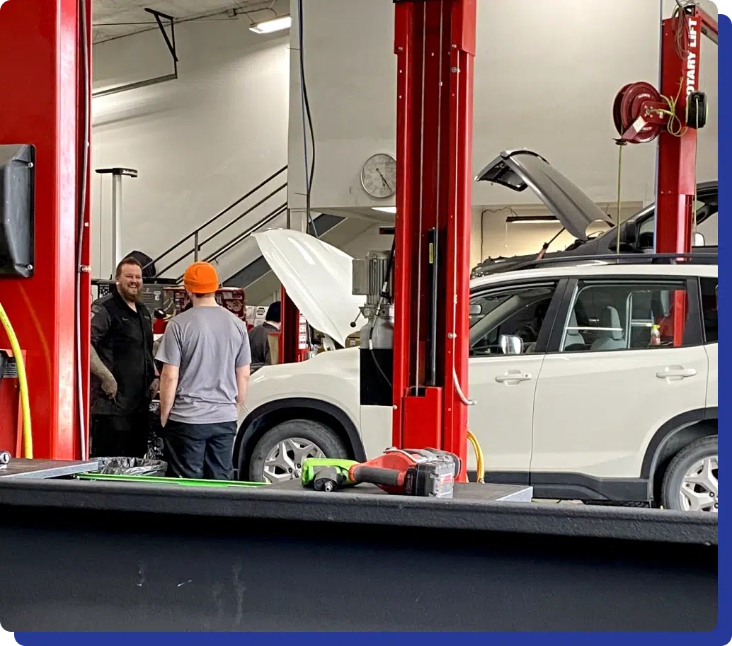 About us Midtown Auto Repair Services in Kodiak, AK. Mechanics conversing in an auto repair shop with a white SUV on a lift, hood open for inspection. Tools in the foreground.