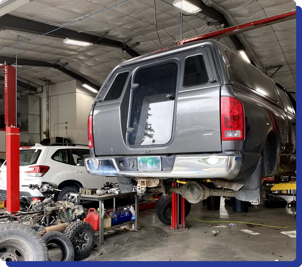 Gray pickup truck on a hydraulic lift in an auto repair shop with another white SUV visible in the background.