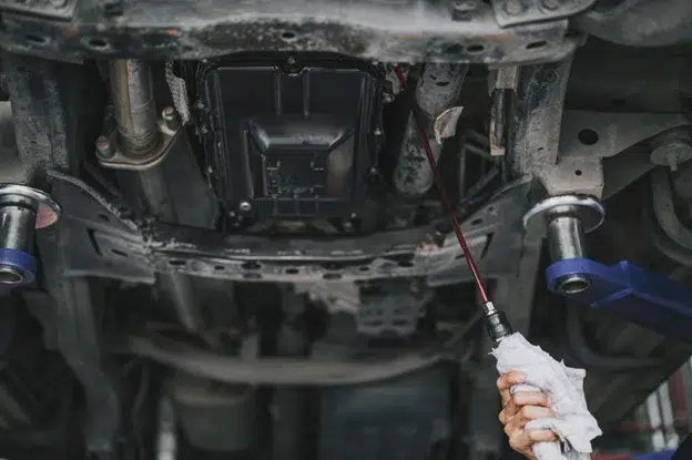 How to spot and address transmission fluid leaks in your vehicle in Kodiak, Ak with Midtown Auto Repair Services. Image of underneath car on lift in the shop with mechanic hand using tool to drain transmission fluid on car while holding a rag around it.